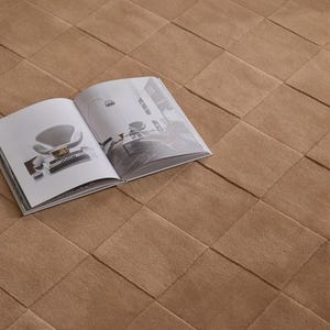 May include: A tan area rug featuring a grid of square tufted sections. An open book rests on the surface, displaying black and white interior design photography and the text Moooi printed on the spine of a book within the image.