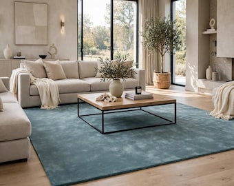 May include: A large, rectangular area rug in a solid, muted teal-blue color with a soft, textured finish. The rug is positioned on a light-toned wooden floor, extending beneath a square wooden coffee table with a black metal frame.