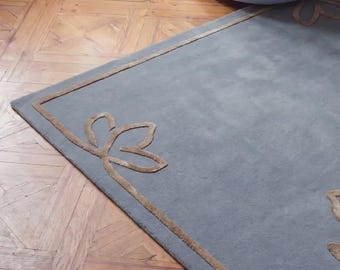 May include: A rectangular area rug in a muted blue-gray tone featuring a raised, tan-colored border and stylized floral corner motifs. The rug has a soft, uniform texture and is positioned on a wooden floor with a herringbone pattern.