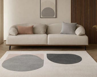 May include: A modern living room scene featuring a cream-colored sofa with two throw pillows and a patterned rug. The rug has gray and pink geometric shapes. A framed abstract art piece hangs on the wall.