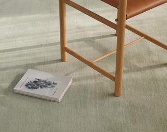 May include: A light green rug with a wooden chair and a book. The chair has a brown leather seat and back. The book has a black and white image on the cover. The chair and book are on the rug.