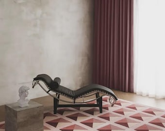 May include: A black leather chaise lounge with a chrome frame, a gray cube side table topped with a white bust sculpture, and a rectangular area rug featuring a geometric pattern of triangles in shades of pink, burgundy, and cream.