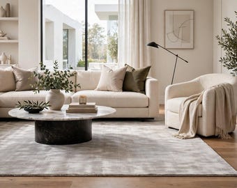 May include: A living room set featuring a cream-colored sectional sofa with beige and olive throw pillows, a round coffee table with a white marble top and a dark cylindrical base, and a cream-colored armchair draped with a beige throw blanket.