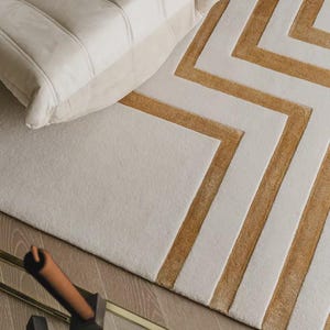 May include: A rectangular area rug featuring a geometric pattern of alternating cream and mustard yellow stripes. The rug has a plush, textured surface with raised lines forming angular shapes. A portion of a cream-colored tufted chair is visible.