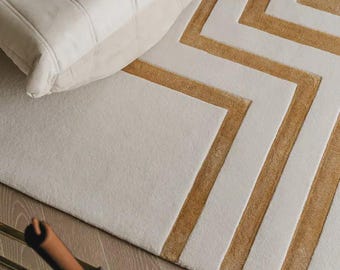 May include: A rectangular area rug featuring a geometric pattern of alternating cream and mustard yellow stripes. The rug has a plush, textured surface with raised lines forming angular shapes. A portion of a cream-colored tufted chair is visible.