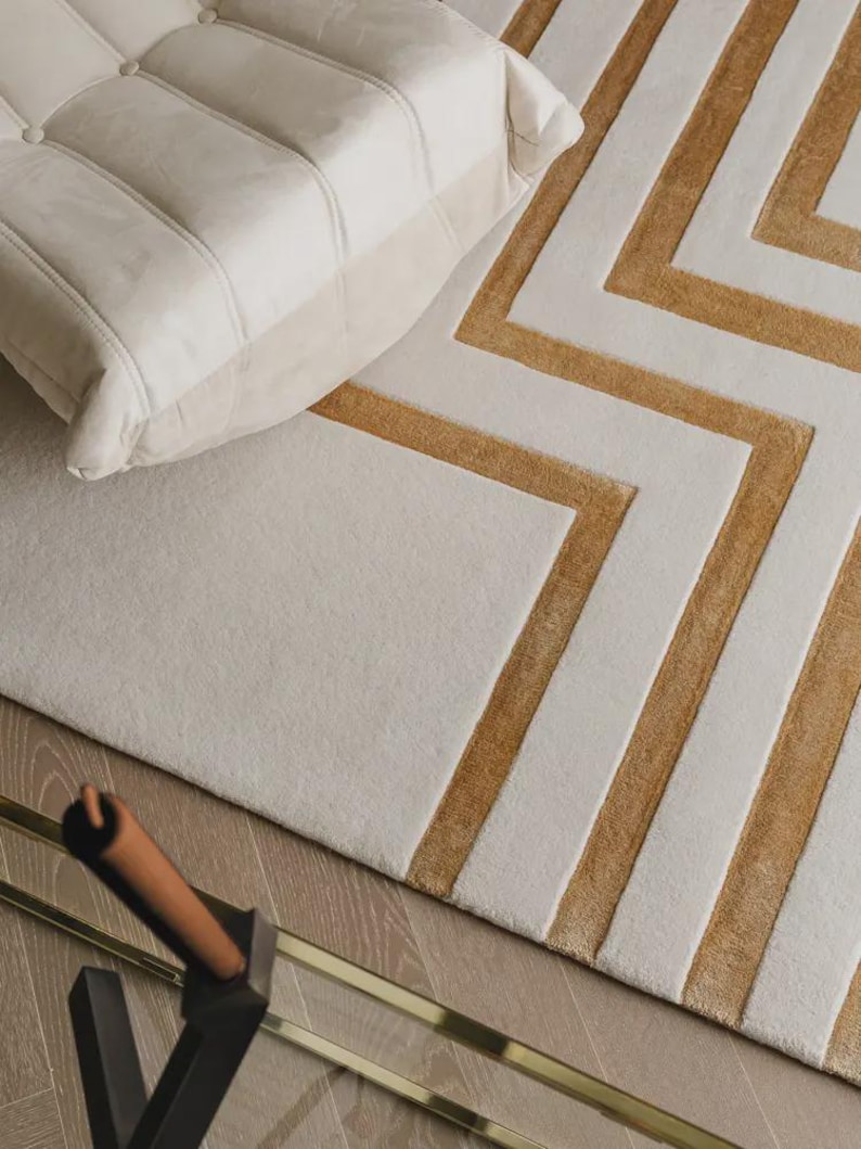 May include: A rectangular area rug featuring a geometric pattern of alternating cream and mustard yellow stripes. The rug has a plush, textured surface with raised lines forming angular shapes. A portion of a cream-colored tufted chair is visible.
