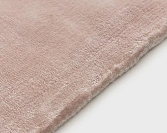 May include: A soft, plush rug in a muted dusty rose color. The surface features a dense, short-pile texture that creates subtle variations in light and shadow across the fabric. The edge of the rug is finished with a clean, straight hem.