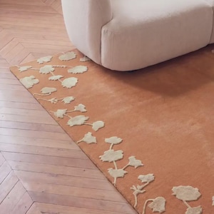 May include: A rectangular area rug featuring a solid terracotta orange base color with a cream-colored floral pattern along the left edge. The rug has a tufted texture and is positioned on a wooden floor next to a curved, cream-colored sofa.