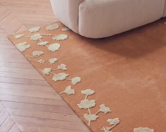 May include: A rectangular area rug featuring a solid terracotta orange base color with a cream-colored floral pattern along the left edge. The rug has a tufted texture and is positioned on a wooden floor next to a curved, cream-colored sofa.