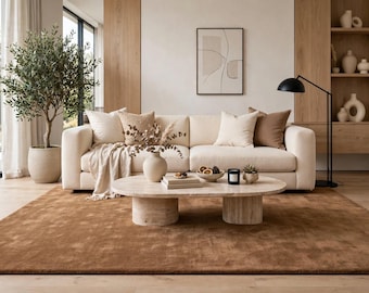May include: A cream-colored sofa with beige and brown throw pillows, a beige throw blanket, and a travertine coffee table. On the table are a vase with dried branches, books, a bowl of dried fruit, a candle, and a small succulent plant.