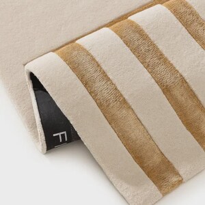 May include: A rectangular rug featuring a cream-colored surface with thick, horizontal golden-brown stripes. The rug is folded over at one corner, revealing a black backing with a partial white letter F visible on the underside of the fabric.