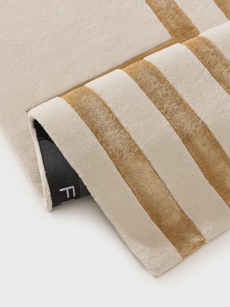 May include: A rectangular rug featuring a cream-colored surface with thick, horizontal golden-brown stripes. The rug is folded over at one corner, revealing a black backing with a partial white letter F visible on the underside of the fabric.