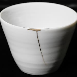 May include: White ceramic cup with a crack repaired with gold-coloured lacquer. The cup has a wide opening and a slightly tapered shape. The repair line runs vertically down the side, creating a unique aesthetic. The cup is set against a black background.