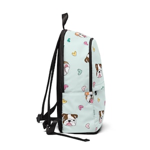 May include: A light blue backpack featuring a repeating pattern of bulldog faces and hearts in pink, yellow, and teal. The backpack has black straps and a top handle. The design is playful and suitable for school or travel.