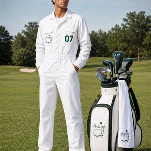 May include: A white jumpsuit with a green Masters logo and the number 07 on the chest, paired with a green Caddies cap and white sneakers. Beside it stands a white and green golf bag containing clubs, a white towel, a golf glove, and two balls.