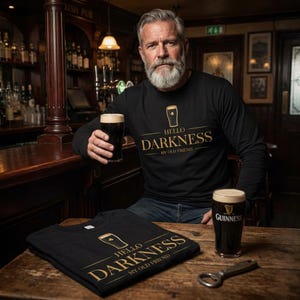 May include: Black long-sleeve shirt with the text "Hello Darkness My Old Friend" in gold. The shirt features a gold graphic of a pint glass. A pint of Guinness and an opener are on the wooden table.