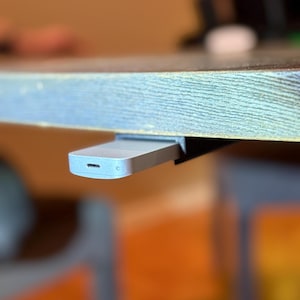 Puede incluir: Una pequeña unidad USB-C plateada está parcialmente insertada en una ranura negra, aparentemente montada debajo de una superficie de madera. La unidad tiene una forma rectangular lisa con esquinas redondeadas.