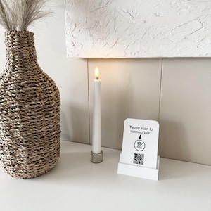 以下が含まれることがあります： QRコードとNFCアイコンが描かれた白いWiFi接続用サイン。Tap or scan to connect WiFiという文字が記載されています。隣には銀色のホルダーに入った白いテーパーキャンドルと、ドライパンパスグラスが入った背の高い編み込みシーグラス製の花瓶が置かれています。