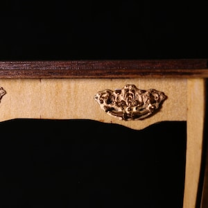 Może przedstawiać: Zbliżenie miniaturowego drewnianego stołu z ciemnobrązowym blatem i jasną drewnianą ramą. Stół posiada dwa ozdobne, złote uchwyty szuflad. Projekt przypomina antyczne meble.