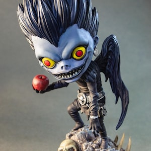 Op de afbeelding: Gedetailleerd beeldje van een personage met een blauwe huid, stekelig zwart haar en grote gele ogen met rode pupillen. Het beeldje houdt een rode appel vast en staat op een voetstuk met schedels en rotsen. Het personage draagt zwarte kleding en heeft zwarte vleugels.