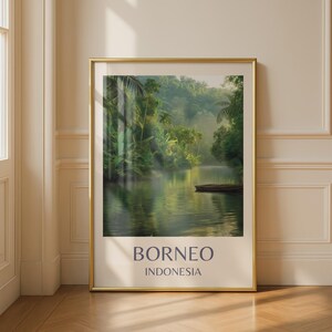 Op de afbeelding: Een ingelijste print met een serene rivierscène in Borneo, Indonesië. Weelderig groen gebladerte omzoomt de oevers, met een kleine boot zichtbaar. De print heeft een gouden lijst en de tekst "BORNEO INDONESIA" in donkerblauw.