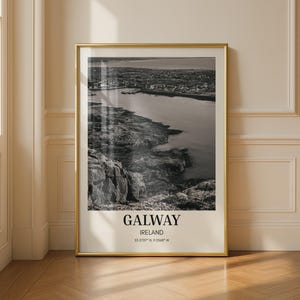 Könnte beinhalten: Ein vertikaler Wandkunstdruck in einem goldenen Rahmen mit einer schwarz-weißen Luftaufnahme der Küste von Galway in Irland, wobei der Text GALWAY, IRELAND und die Koordinaten 53.2707° N, 9.0568° W unten aufgedruckt sind.