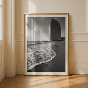 Könnte beinhalten: Ein schwarz-weiß gerahmter Wandkunstdruck, der einen hohen, geschwungenen Wolkenkratzer an einem Sandstrand zeigt. Die Meereswellen spülen sanft an das Ufer im Vordergrund und erzeugen einen Kontrast zwischen dem dunklen Sand und dem weißen Meeresschaum.