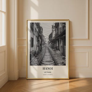 Könnte beinhalten: Schwarz-Weiß-Druck in einem goldenen Rahmen, der eine Bahngleisstrecke zwischen Gebäuden zeigt. Der Druck enthält den Text "HANOI VIETNAM" und Koordinaten.