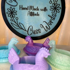 Puede incluir: Una colecci&oacute;n de recipientes con forma de gallina en tonos azules, morados y verdes. Los recipientes est&aacute;n frente a un letrero redondo con las palabras "Hand Made with Attitude Cuss Words".