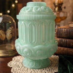 May include: A mint green decorative jar with a lid, featuring intricate floral and arched patterns. The jar sits on a cream-colored, crocheted doily. The jar is approximately 8 inches tall.