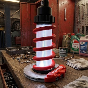 May include: A unique lamp with a red coil design, a black top, and a silver base, resembling a car suspension. The lamp is on a wooden workbench with tools and automotive parts, including a red Brembo brake caliper.