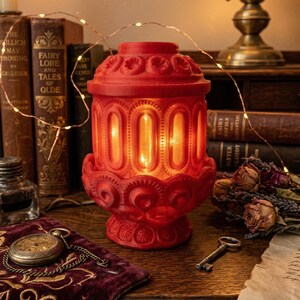 May include: A red, ornate lantern with a decorative base and top, featuring vertical oval windows. The lantern is lit from within, casting a warm glow. The image includes antique books, a pocket watch, and a key, suggesting a vintage or gothic theme.