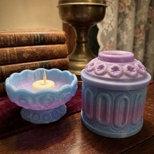 May include: Two decorative candle holders in blue and purple hues. One is a pedestal bowl with a lit candle, the other is a small, lidded container. Both have intricate, embossed patterns. The items are on a wooden surface, with a vintage lamp and books in the background.