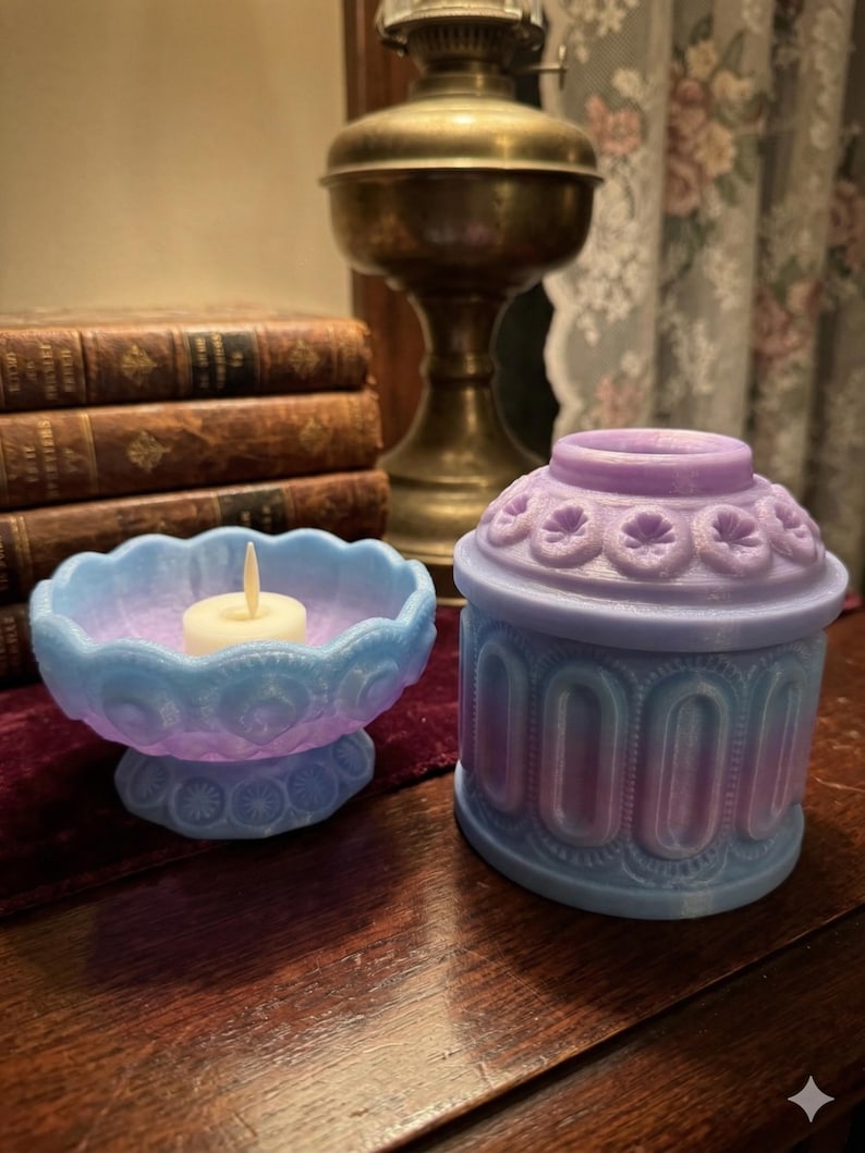May include: Two decorative candle holders in blue and purple hues. One is a pedestal bowl with a lit candle, the other is a small, lidded container. Both have intricate, embossed patterns. The items are on a wooden surface, with a vintage lamp and books in the background.