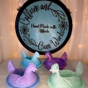 Puede incluir: Cuatro macetas con forma de gallina en tonos azules, morados y verdes. Un letrero redondo con las palabras "Caffeine and Cuss Words" y "Hand Made with Attitude" cuelga sobre las macetas. El letrero es negro con el centro azul claro.