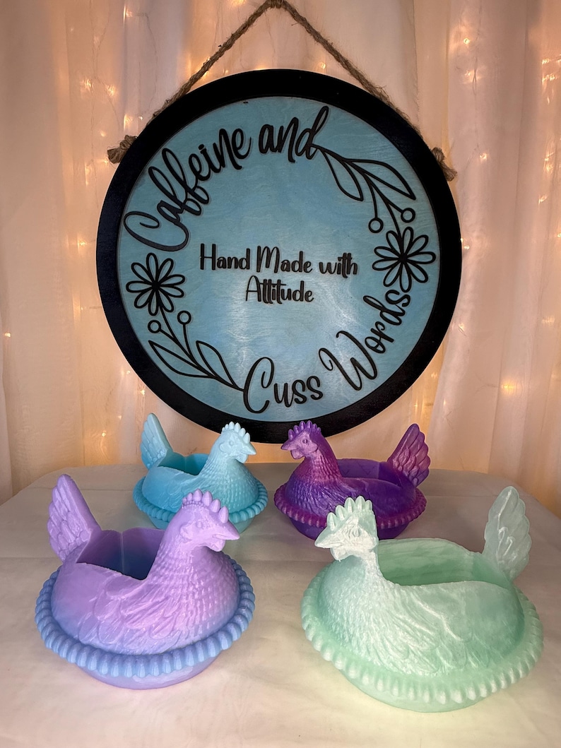 Puede incluir: Cuatro macetas con forma de gallina en tonos azules, morados y verdes. Un letrero redondo con las palabras "Caffeine and Cuss Words" y "Hand Made with Attitude" cuelga sobre las macetas. El letrero es negro con el centro azul claro.