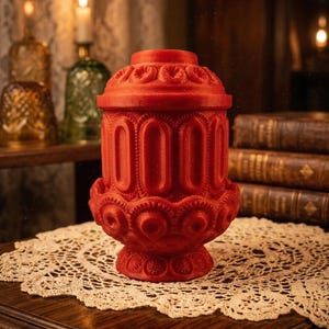 May include: A red, ornate candle holder with a lid, featuring a detailed, repeating pattern of arches and floral designs. The candle holder is placed on a lace doily, with a blurred background of candles and books.