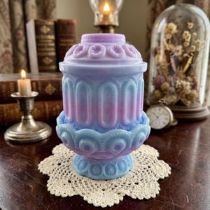 May include: A pastel-colored, antique-style jar with a lid, featuring a blend of blue, pink, and purple hues. The jar has a decorative, ornate design and sits on a cream-colored, crocheted doily. A vintage aesthetic.