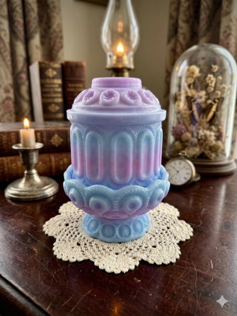 May include: A pastel-colored, antique-style jar with a lid, featuring a blend of blue, pink, and purple hues. The jar has a decorative, ornate design and sits on a cream-colored, crocheted doily. A vintage aesthetic.