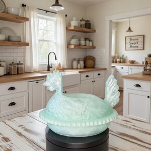 Puede incluir: Un plato decorativo en forma de gallina azul claro, posiblemente para servir, se encuentra sobre una base giratoria negra. El plato est&aacute; sobre una mesa de madera blanca desgastada en un entorno de cocina. La cocina tiene armarios blancos y estantes de madera.