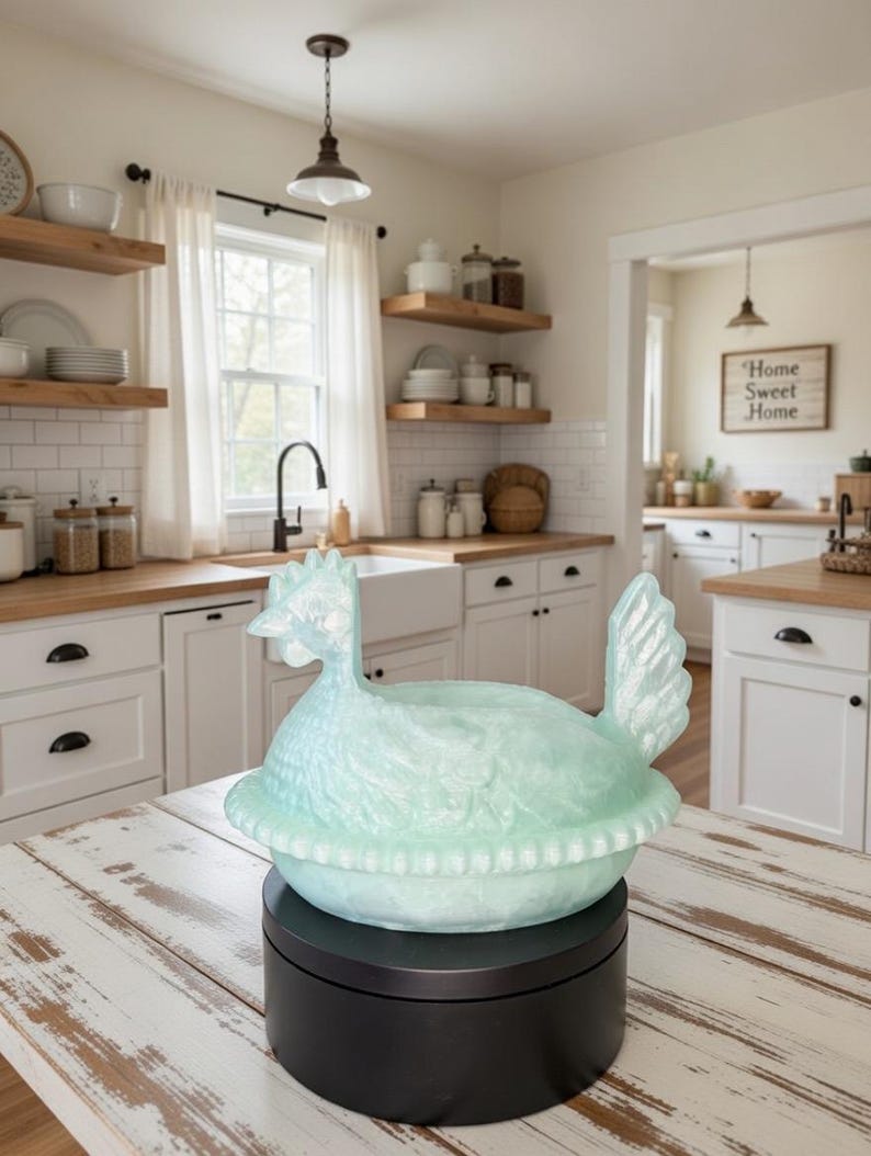 Puede incluir: Un plato decorativo en forma de gallina azul claro, posiblemente para servir, se encuentra sobre una base giratoria negra. El plato est&aacute; sobre una mesa de madera blanca desgastada en un entorno de cocina. La cocina tiene armarios blancos y estantes de madera.