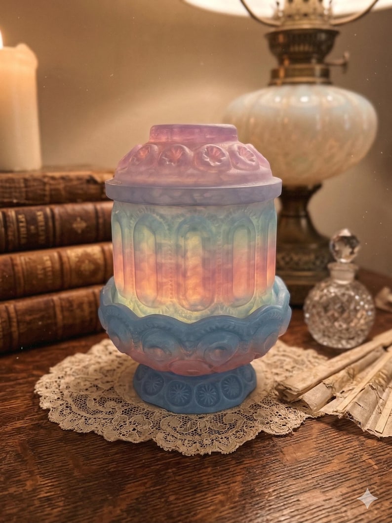 May include: A pastel-colored decorative glass lantern with a lid, featuring a gradient of pink, blue, and yellow hues. The lantern has a detailed, ornate design and sits on a decorative base. The image is set on a wooden surface with antique books and a lace doily.