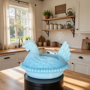 Puede incluir: Un plato azul claro con forma de gallina, probablemente para servir o decorar, est&aacute; sobre un soporte giratorio negro. El plato est&aacute; en un entorno de cocina con encimeras de madera, armarios blancos y una ventana.