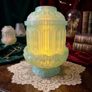 May include: A light blue, ornate glass lantern with a warm yellow glow. The lantern features a detailed architectural design and sits on a matching base. It is placed on a white lace doily, suggesting a vintage or antique aesthetic.