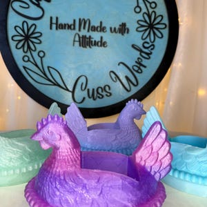 Puede incluir: Una colecci&oacute;n de macetas con forma de gallina en tonos morado, azul y verde. Las macetas tienen texturas detalladas de plumas y crestas. Un letrero redondo en el fondo dice "Caffeine and Cuss Words" y "Hand Made with Attitude."