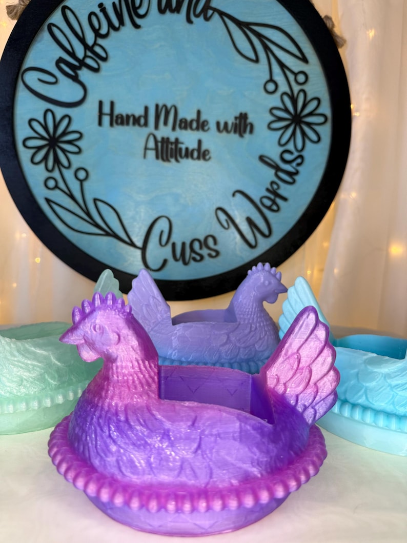 Puede incluir: Una colecci&oacute;n de macetas con forma de gallina en tonos morado, azul y verde. Las macetas tienen texturas detalladas de plumas y crestas. Un letrero redondo en el fondo dice "Caffeine and Cuss Words" y "Hand Made with Attitude."