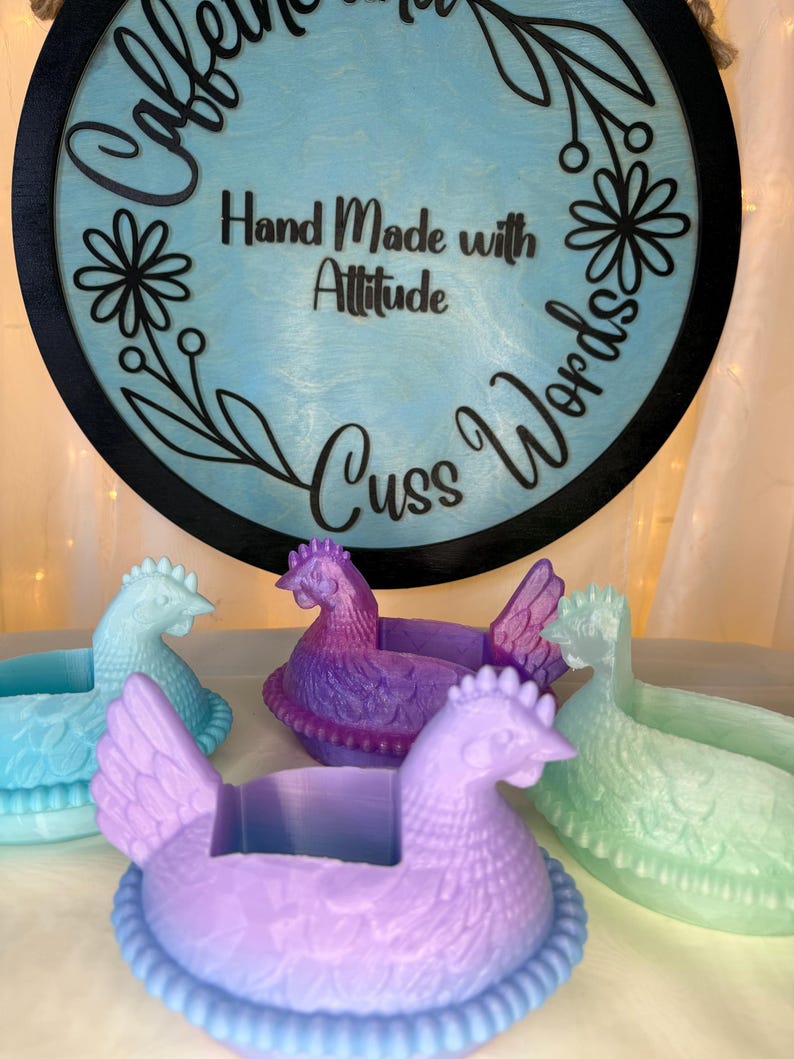 Puede incluir: Una colecci&oacute;n de recipientes con forma de gallina en tonos azules, morados y verdes. Los recipientes est&aacute;n frente a un letrero redondo con las palabras "Hand Made with Attitude Cuss Words".