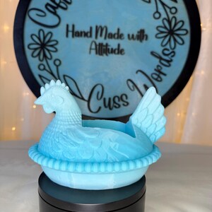 Puede incluir: Un recipiente decorativo en forma de gallina azul claro. La gallina est&aacute; sentada sobre una base en forma de nido. El fondo presenta un letrero redondo con las palabras "Caffeine and Cuss Words" y "Hand Made with Attitude".