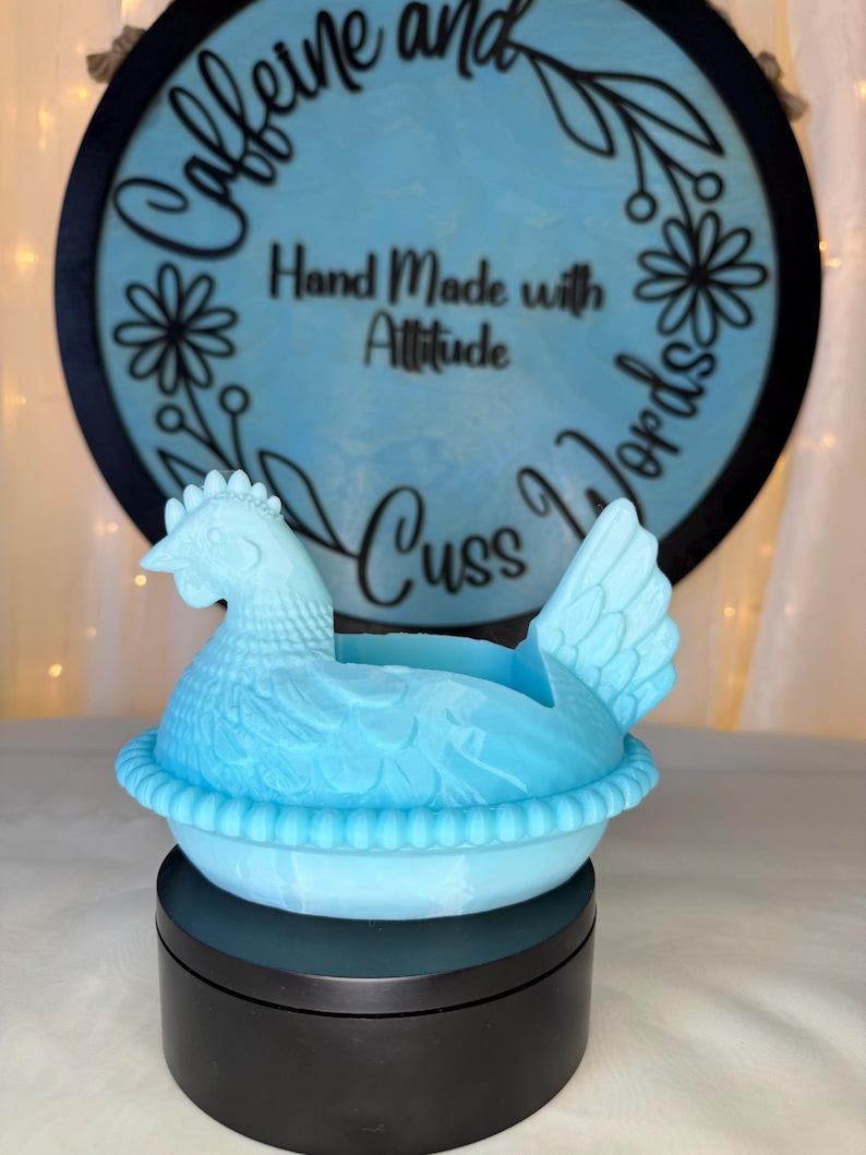 Puede incluir: Un recipiente decorativo en forma de gallina azul claro. La gallina est&aacute; sentada sobre una base en forma de nido. El fondo presenta un letrero redondo con las palabras "Caffeine and Cuss Words" y "Hand Made with Attitude".