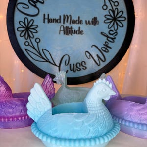 Puede incluir: Un recipiente en forma de gallina azul claro con un dise&ntilde;o detallado, sentado en una base en forma de nido. La imagen tambi&eacute;n muestra otros recipientes en forma de gallina en p&uacute;rpura y verde. Un letrero de madera en el fondo dice "Caffeine and Cuss Words" y "Hand Made with Attitude."